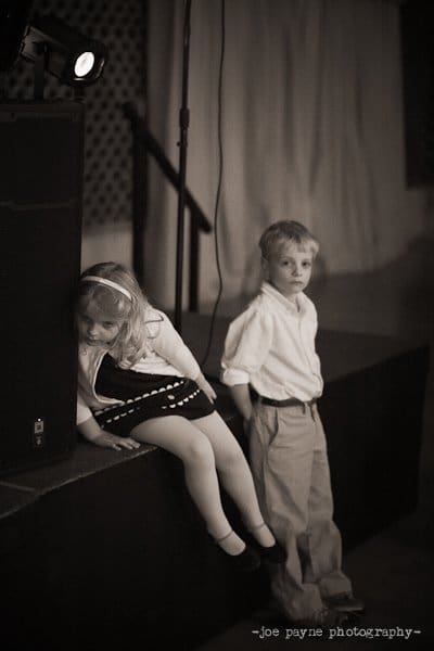 A black and white photo of a young girl and boy. The girl leans against a speaker with a tired expression, wearing a headband and a dress. The boy stands beside her with his hands in his pockets, dressed in a shirt and trousers.