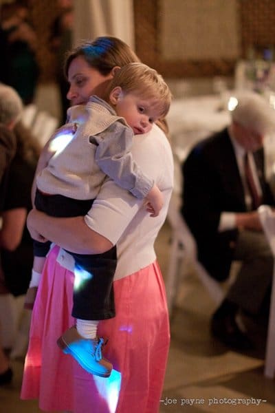 A woman holds a young child in her arms. The child is wearing a vest, shirt, and blue shoes. They appear to be in a warmly lit room with other people sitting at tables in the background.