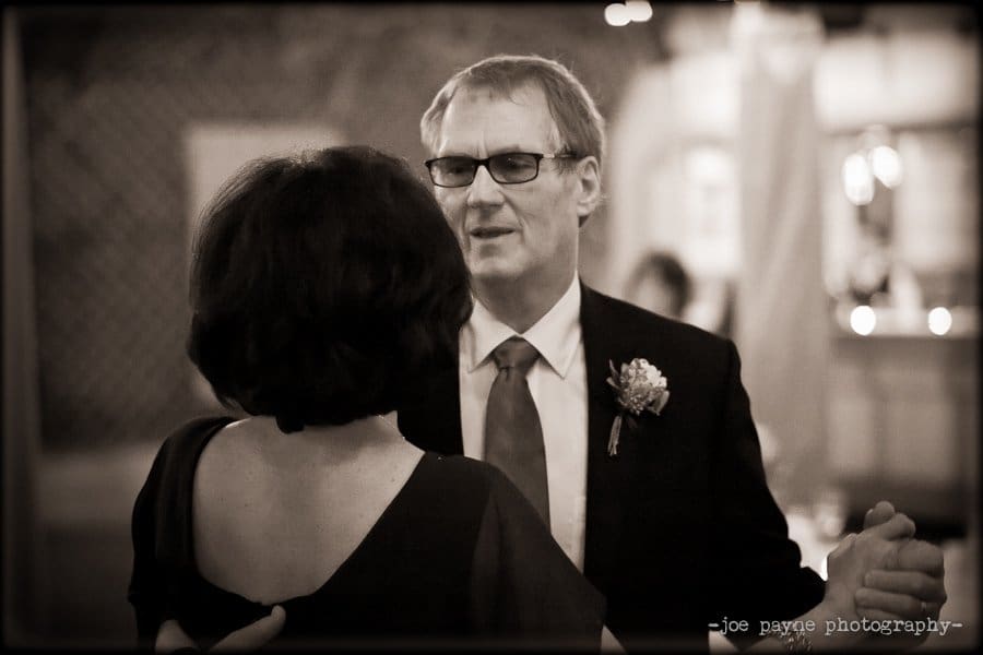 A man in a suit with glasses dances with a woman. The atmosphere is warm and intimate, with soft lighting in the background. The photo is in black and white, giving it a timeless feel.