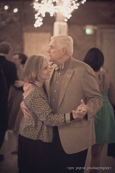 An elderly couple dances closely, surrounded by softly lit decorations at what appears to be a formal event. The atmosphere is warm and intimate, and they are dressed in elegant attire.