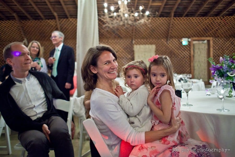 A woman smiling and seated, holding two young girls on her lap, at a party. One girl wears a floral dress and the other is in a cozy sweater. Guests are in the background, and a chandelier hangs from the ceiling. Tables are adorned with purple flowers.