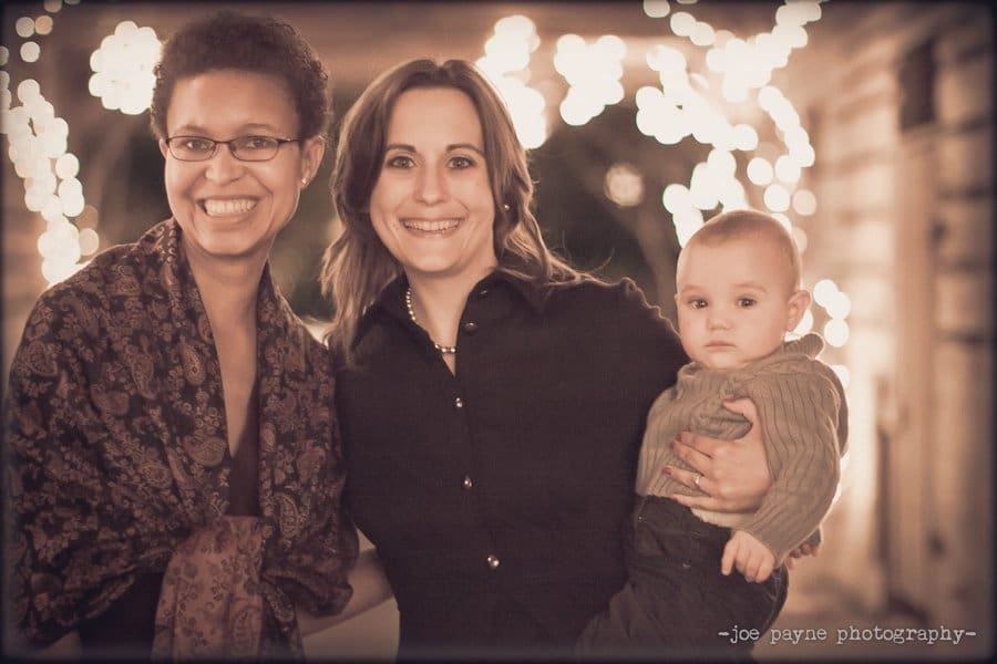 Two women smiling, standing under string lights. One woman holds a baby. The photo has a warm, soft glow.