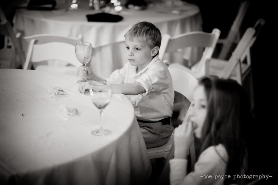 A young boy sits at a round table, holding a wine glass and looking ahead thoughtfully. A young girl with long hair sits beside him, resting her hand near her mouth. The setting suggests a formal event.