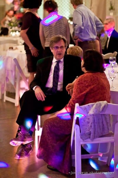 A man in a suit and tie sits across from a woman in a patterned dress at a wedding reception. Colorful lights reflect on the floor and walls, creating a festive atmosphere. Other guests are visible in the background.