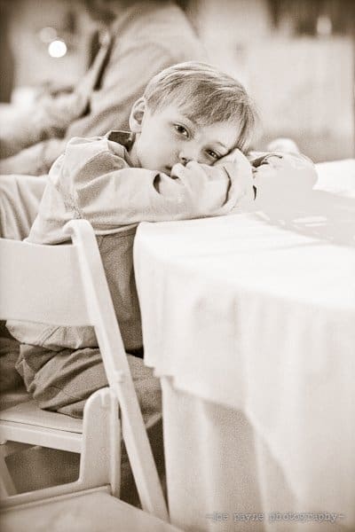 A young child with a thoughtful expression sits at a table, resting their head on their arms. The image is in black and white, with a soft, nostalgic atmosphere.
