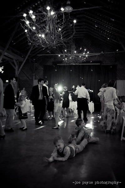 Black and white photo of a lively wedding reception in a large hall with chandeliers. Guests are dancing, while two children play on the floor in the foreground. The atmosphere is joyful and festive.