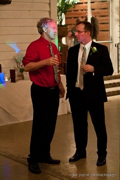 Two men in formal attire are standing and conversing. One is wearing a red shirt with a patterned tie, and the other is in a suit with a boutonniere. Both are holding drinks, and they appear to be at a social event or celebration indoors.