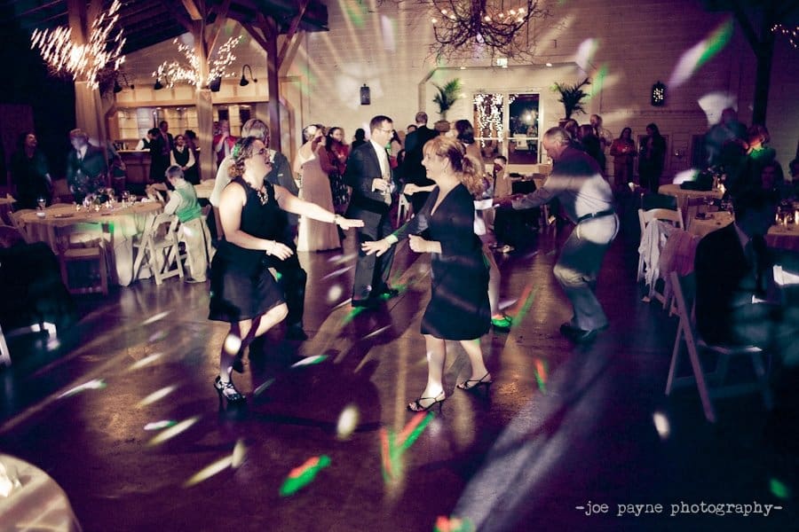 People dancing joyfully in a warmly lit indoor venue with colorful lights scattered across the floor. Tables and chairs are visible in the background, and some people are seated, enjoying the festive atmosphere.