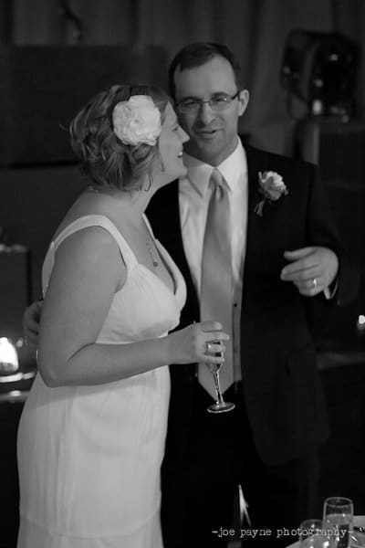 A couple, dressed in formal attire, stands together in a dimly lit room. The woman wears a white dress with a flower in her hair, holding a champagne flute. The man is in a suit with a boutonniere, engaging in conversation.