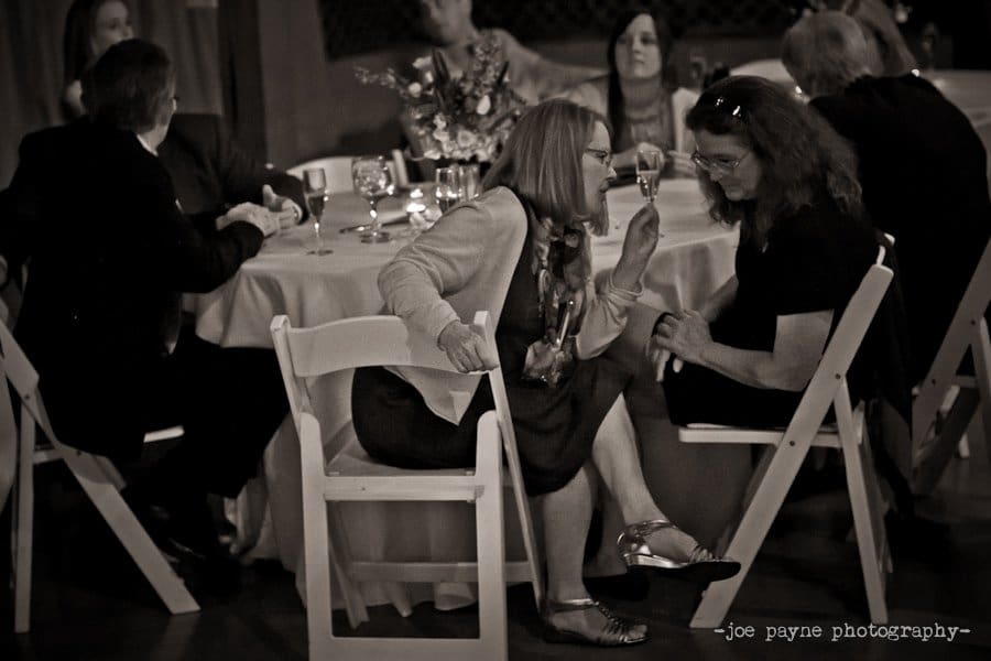 A black and white photo of people sitting around a table at an event. Two women in the foreground are talking, while others in the background are engaged in conversation. The table has glasses and a flower centerpiece.