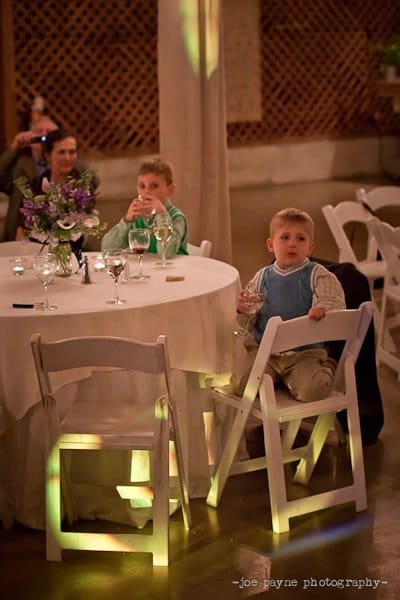 Two young boys sit at a round table with a white tablecloth and glassware in a dimly lit room. A person in the background holds a camera. A floral centerpiece is on the table. The scene has soft, colorful lighting effects.