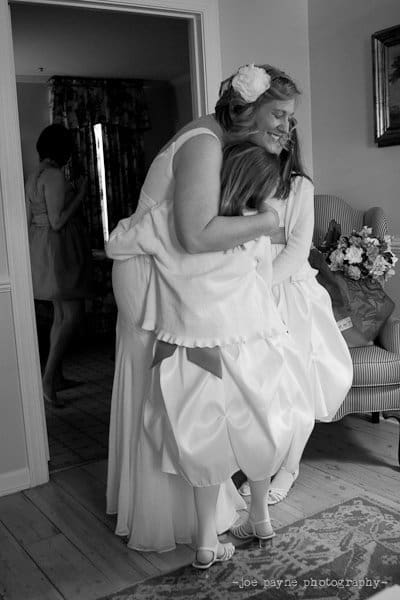 A bride in a white dress hugs two young girls in matching white dresses. They are inside a room with a patterned armchair and a bouquet of flowers nearby. Another woman stands in the background. The scene appears joyful and intimate.