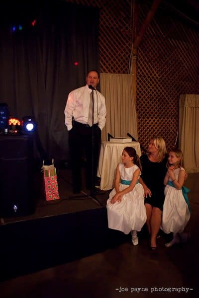 A man in a white shirt and tie stands at a microphone on a stage. A woman in a black dress sits nearby with two young girls in white dresses listening attentively. The background features dark curtains and lattice walls.