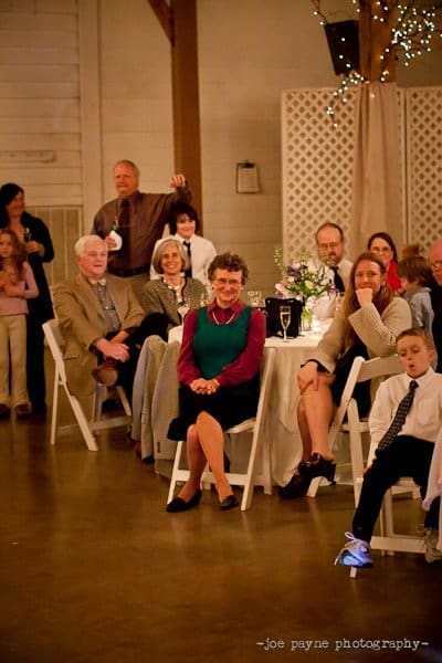 A group of people sit around a table, attentively watching an event. A child near the front is wearing sneakers with glowing lights. The setting has a cozy, rustic ambiance with soft lighting and greenery decor.
