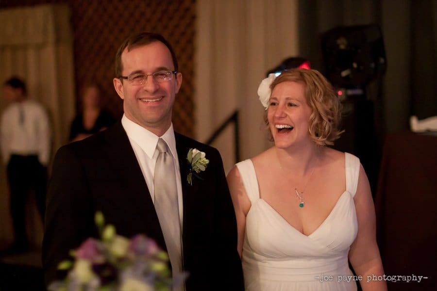 A smiling couple in formal attire stands together. The man wears a dark suit with a white shirt and tie, and the woman is in a white dress with a floral accessory in her hair. They are in a warmly lit indoor setting.