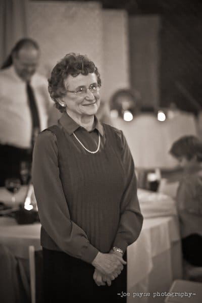An elderly woman stands smiling at an event, wearing glasses and a pearl necklace. The background includes people seated at tables adorned with candles, creating a warm and intimate atmosphere.