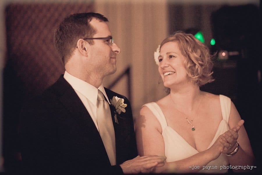 A couple in formal attire share a joyful moment. The man, in a suit and tie, gazes at the woman, who is smiling and wearing a white dress with a flower in her hair. They appear to be at a wedding or formal event.