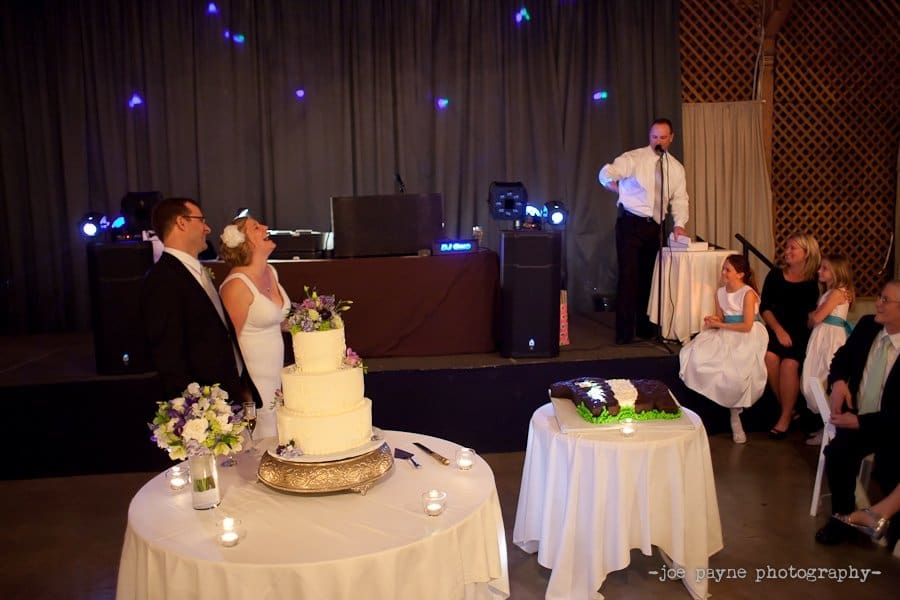 A bride and groom laugh together at their wedding reception. A multi-tiered cake and gift table are in the foreground. A man speaks into a microphone on a small stage, with bridesmaids seated nearby.