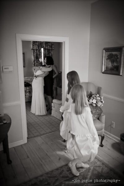 A bride in a wedding dress hugs a woman in a dark dress in a room. Two young girls in dresses approach, smiling. The setting is a cozy home with framed artworks and a vase of flowers. Black and white image.
