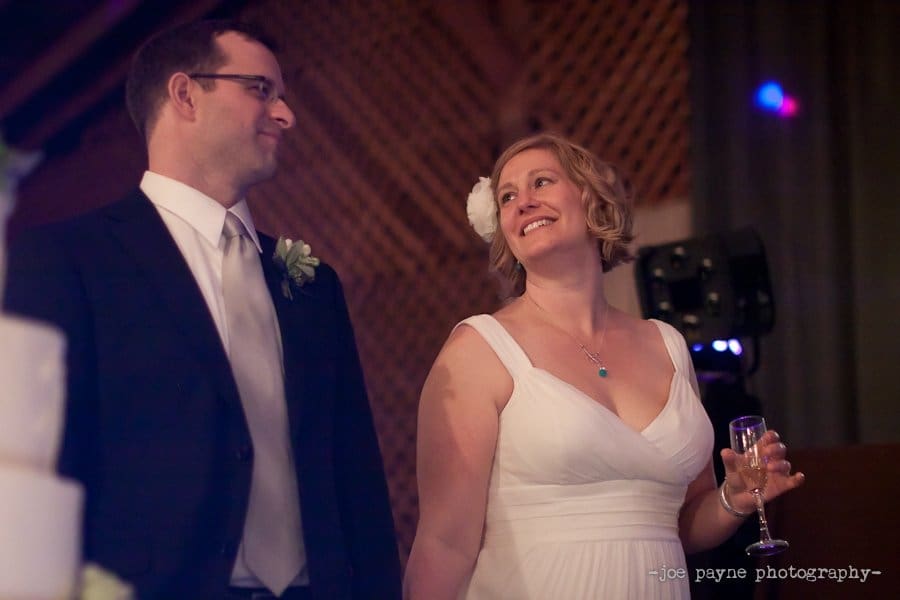 A couple in formal attire smiles at each other, standing beside a multi-tiered wedding cake. The woman holds a champagne glass and wears a white dress with a floral hair accessory. The background is softly lit, creating a warm, intimate atmosphere.