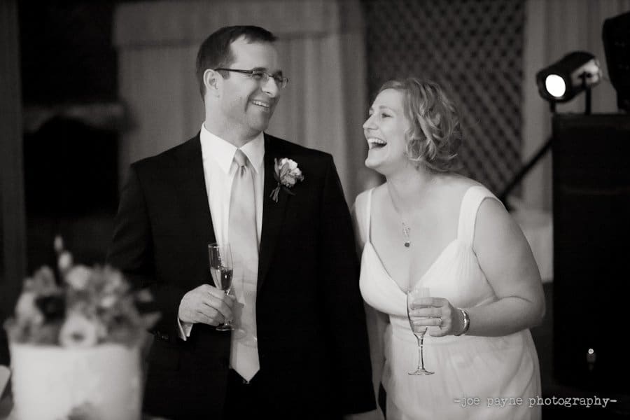 A couple in formal attire is smiling and holding champagne glasses at an event. The woman is laughing joyfully while the man looks at her. A decorated cake is visible in the foreground.
