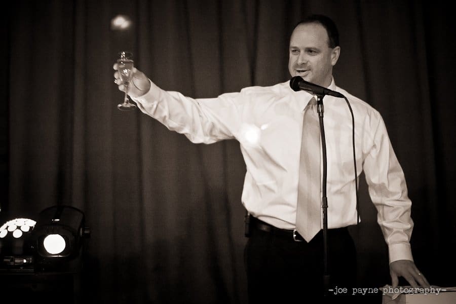 A man in a formal outfit stands holding a microphone and raising a glass in a toast. He smiles as he addresses an audience. The background is a plain, dark curtain, and theres a light source beside him. The image is in black and white.