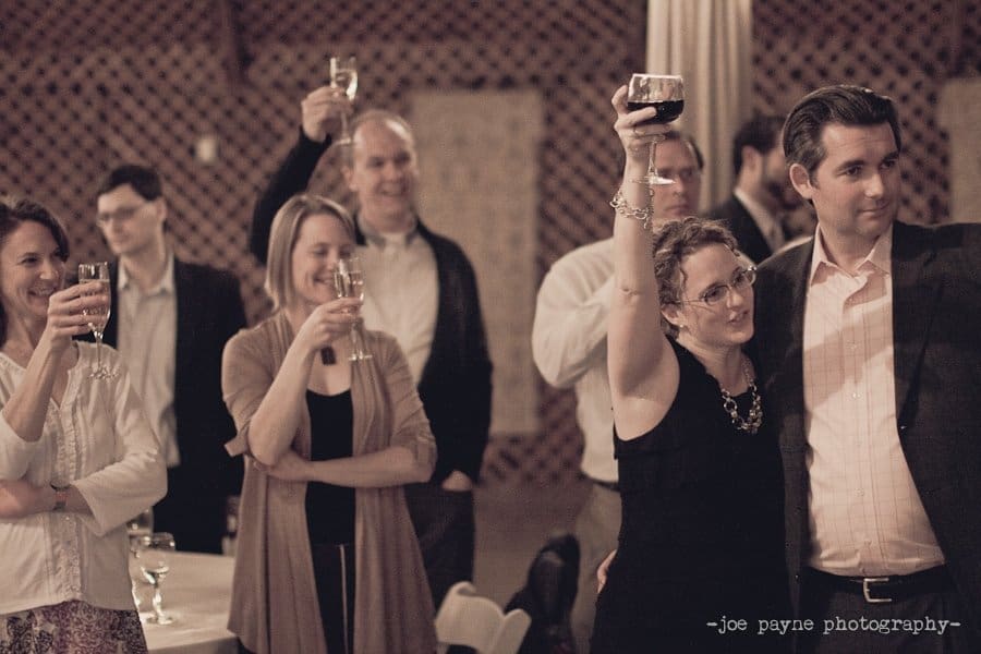 A group of people at a celebration raise their glasses in a toast. The guests are dressed in semi-formal attire and appear joyful. The setting has a warm, inviting atmosphere with soft lighting.