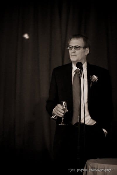 A man in a suit stands holding a glass in front of a microphone, appearing to give a speech. The setting is formal, and the background is dimly lit.