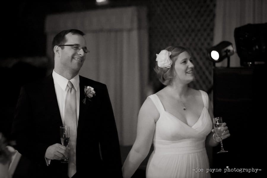 A couple in formal attire holds hands and champagne glasses, smiling and looking to the side. The woman wears a white dress and a flower in her hair, and the man wears a suit with a boutonniere. Its a joyful, intimate moment at an event.