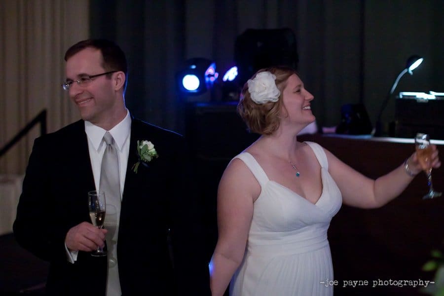 A couple in formal attire, both smiling, hold champagne glasses. The woman wears a white dress with a flower in her hair, and the man is in a suit with a boutonniere. They appear to be celebrating at an indoor event with dim lighting.