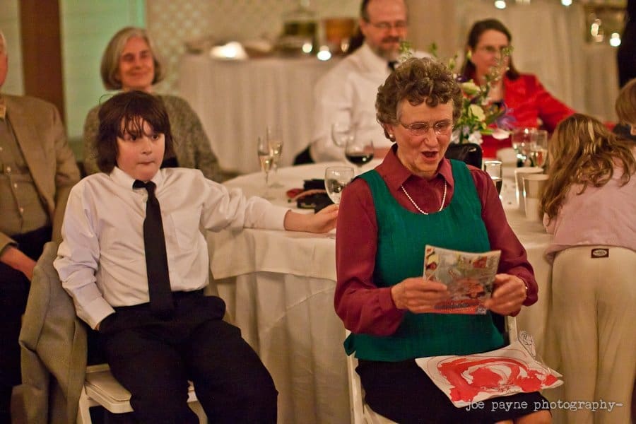An elderly woman in a green vest and red shirt is joyfully reading a colorful card. A young boy in a white shirt and black tie sits beside her, looking amused. Several people are seated at tables in the dimly lit background.