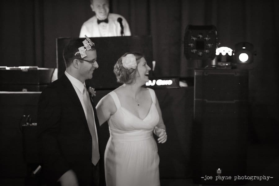 A bride and groom, both wearing playful headbands, smile and dance together at a wedding reception, with a DJ in the background. The image is in black and white.