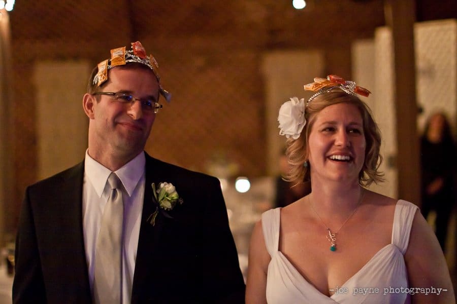 A bride and groom wearing white outfits stand smiling. Both have packets of hot sauce on their heads as playful accessories. The background is softly lit, creating a warm atmosphere.