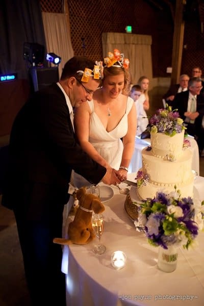 A couple in formal attire cuts a wedding cake. The woman wears a dress with condiment packets in her hair, and the man is in a suit. A stuffed animal and floral arrangement decorate the table. Guests are visible in the background.