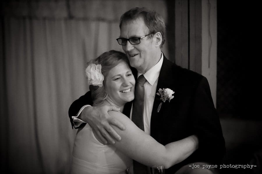 A bride and an older man share a joyful embrace during a wedding. The bride has a flower in her hair and is wearing a white dress, while the man is in a dark suit with a tie and boutonniere. The background is softly lit and out of focus.