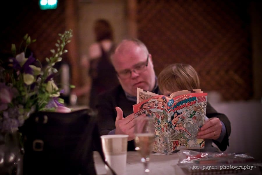 A man and a child sit together at a table, engrossed in reading a comic book. The table is adorned with a flower arrangement, a purse, and a plastic bag. The background is softly blurred, creating a warm and intimate atmosphere.