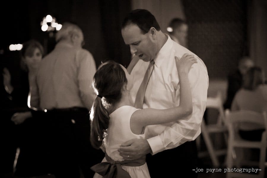 A man in a white shirt dances closely with a young girl in a white dress at a formal event. Other guests can be seen socializing in the dimly lit background.