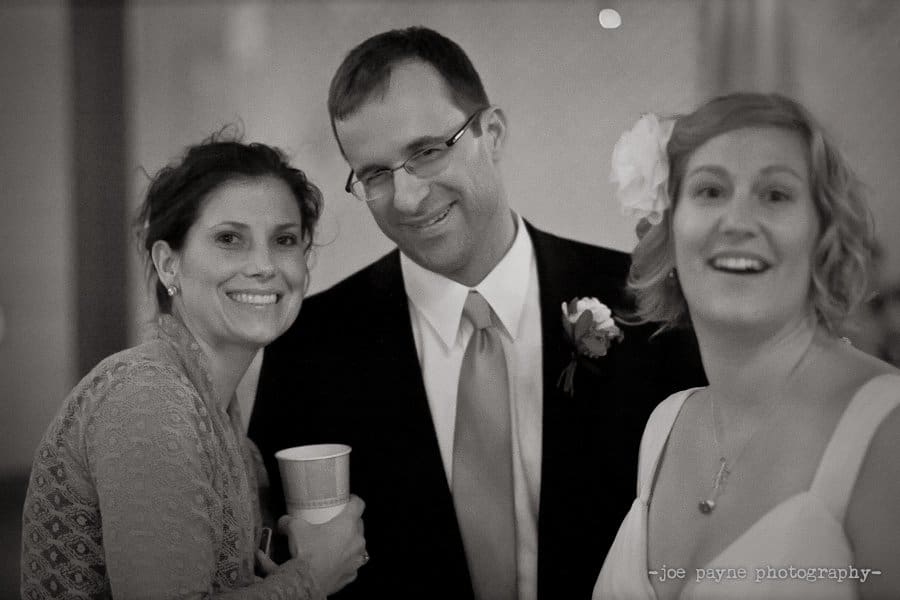 Three people pose for a photo at a formal event. Two women, one with a drink and another with a flower in her hair, stand beside a man in a suit and tie. They are smiling and appear happy. The image is in black and white.