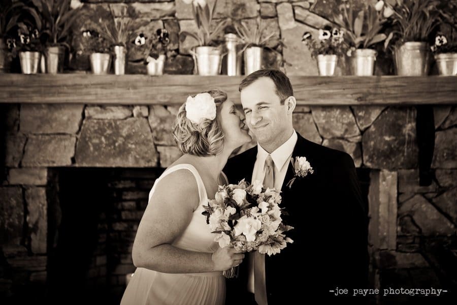 A bride and groom share a sweet moment in front of a rustic stone fireplace lined with potted flowers. She holds a bouquet and smiles towards the camera, while he gently kisses her cheek. The photo captures a joyful wedding scene.