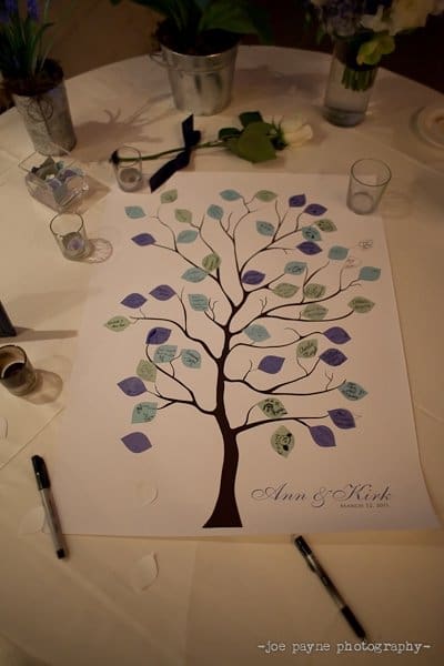 A wedding guestbook poster featuring a tree with colored leaves for guests to sign, displayed on a table with pens and small vases. The names Ann & Kirk and date August 31, 2013 are written at the bottom.