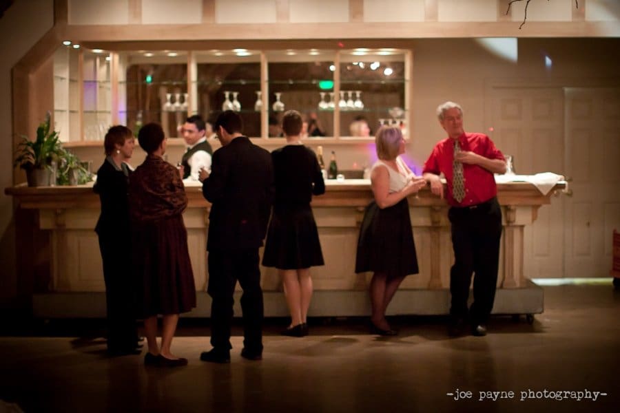 A group of people in formal attire stand and chat around a bar. A bartender serves drinks in the background, while a man in a red shirt leans against the counter. The room is softly lit, creating a warm atmosphere.