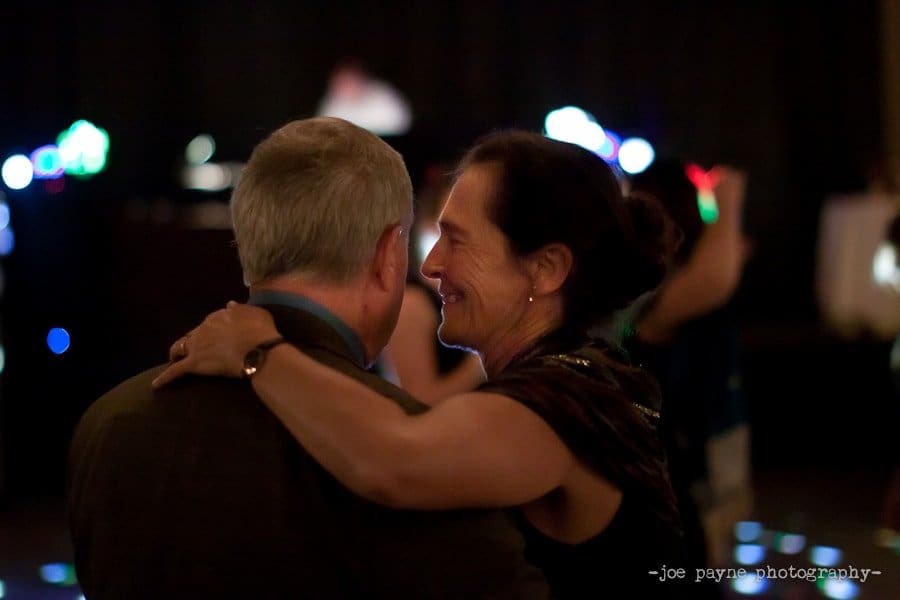 A couple dances closely in a dimly lit room, the woman smiling with her arm around the mans shoulder. Blurred lights in the background create a warm, intimate atmosphere.