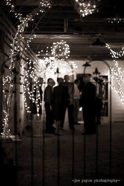 A black and white image of a group of people gathered under a decorated, string-lit porch. The scene looks festive with soft lights illuminating the area, casting gentle shadows. The focus is on the ambient and cozy atmosphere.