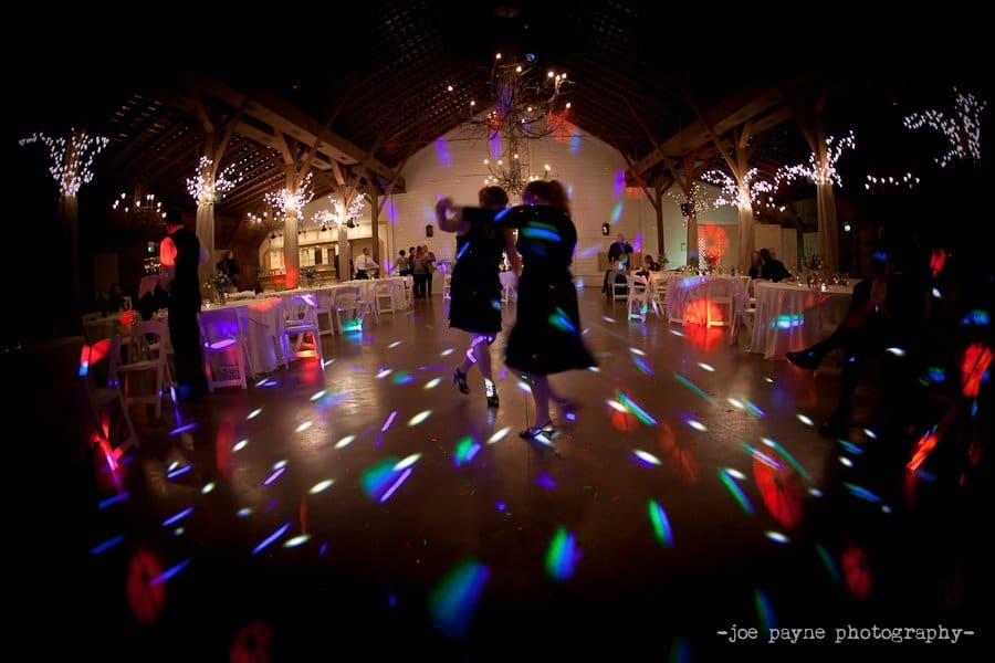 A dimly lit dance floor features two people dancing under colorful disco lights. The space is decorated with string lights and white tables.