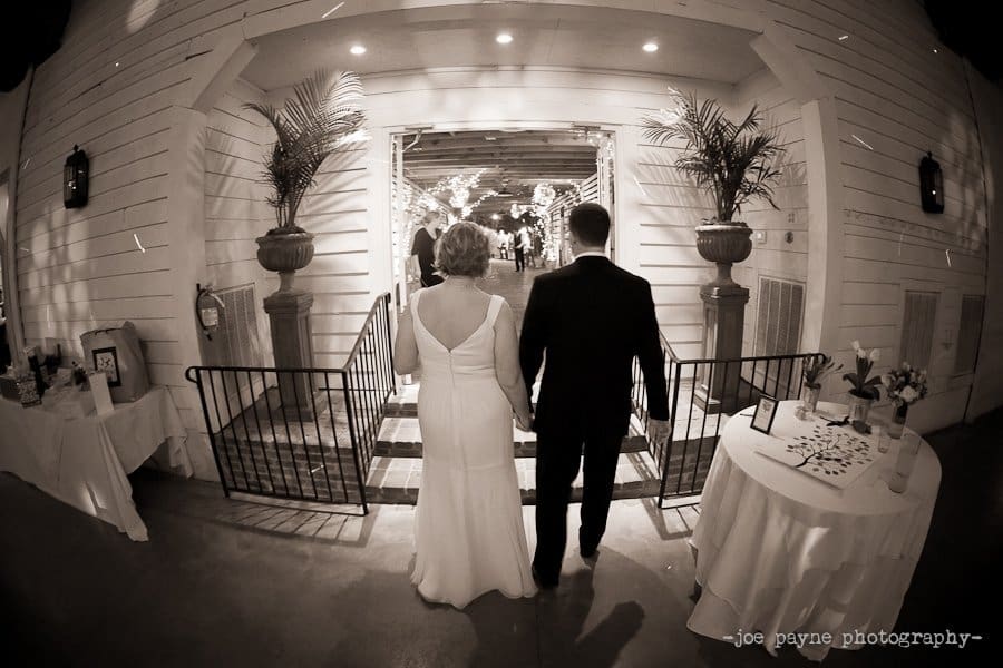 A bride and groom walk hand in hand toward a warmly lit reception area, adorned with potted plants. They are seen from behind, entering through a decorated archway in a spacious venue.