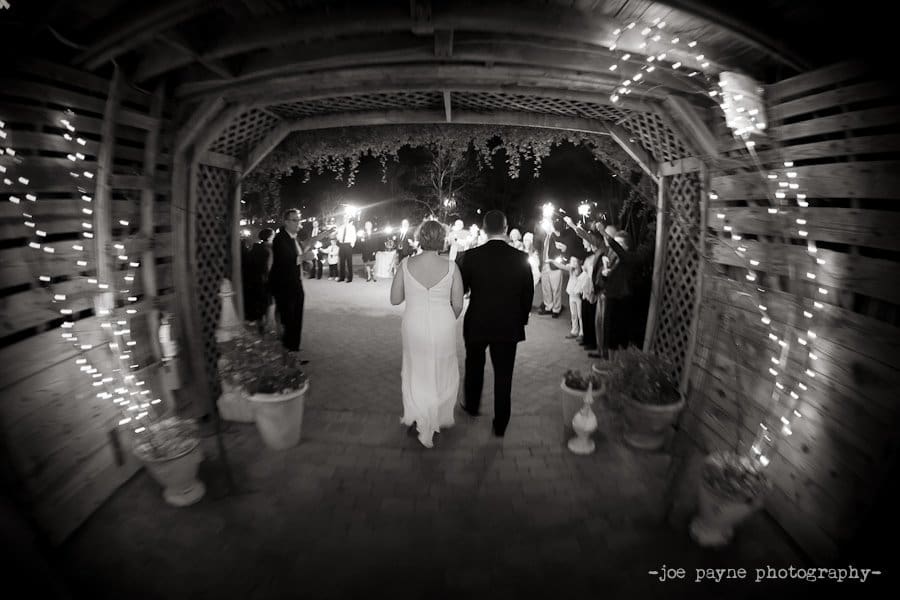 A bride and groom walk hand in hand through a wooden archway adorned with string lights, surrounded by guests holding sparklers in celebration. The scene is captured in black and white, creating an elegant and timeless atmosphere.