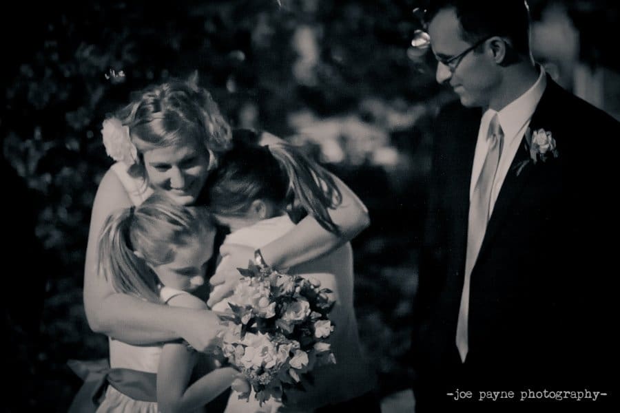 A woman embraces two young girls, all smiling, holding a bouquet. A man in a suit stands beside them, smiling warmly. The photo has a soft, nighttime setting.
