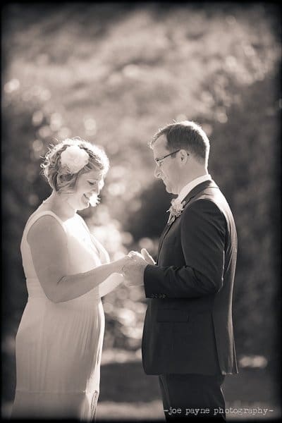 A black and white photo of a bride and groom standing hand in hand outdoors. The bride is wearing a sleeveless dress with a flower in her hair, and the groom is in a suit, both smiling at each other.