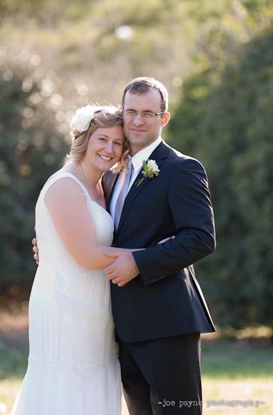 A bride and groom stand outdoors, smiling and embracing each other. The bride is wearing a white dress with a flower in her hair, and the groom is in a dark suit with a boutonniere. The background is lush greenery under a sunny sky.