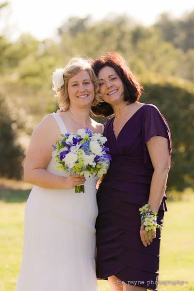 Two women standing together outdoors, smiling. One is holding a bouquet of white and purple flowers, wearing a white dress with a flower in her hair. The other is in a purple dress with a wrist corsage. They appear to be enjoying a sunny day.